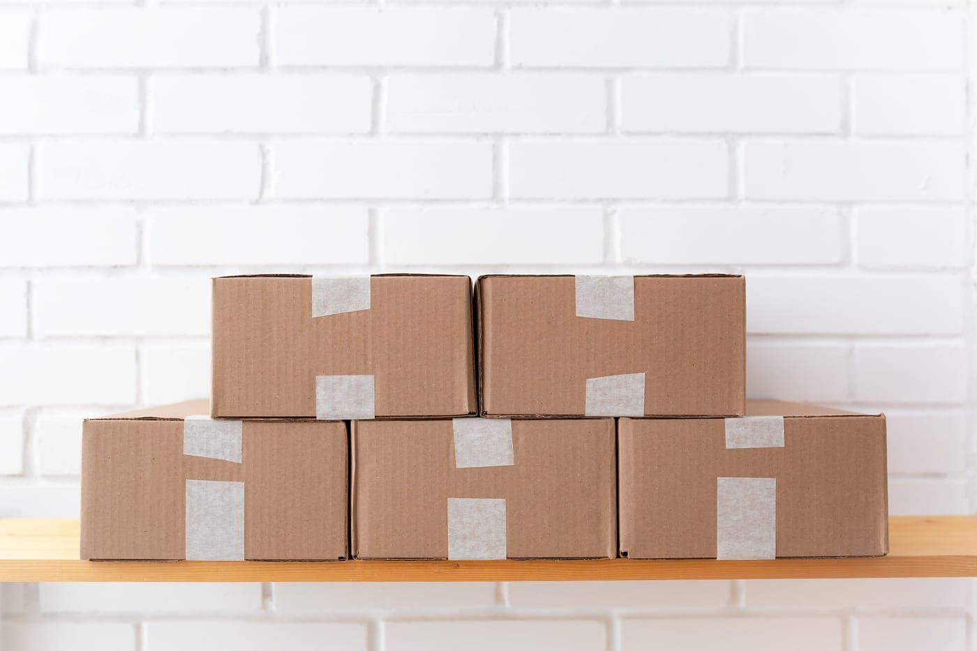 Taped cardboard boxes stacked on a wooden shelf against a white brick wall
