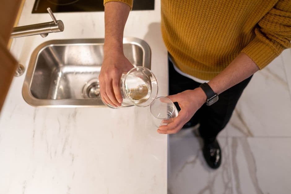 A person pouring water from one glass into another over a kitchen sink